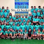 Group photo of Sis-Q Seeker Camp 2024 participants in teal shirts, gathered under a banner indoors smiling.