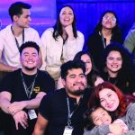 A diverse group of people smiling and sitting together indoors, with a guitar in the background.
