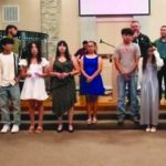 A group of people standing in a church setting, dressed casually, with a stage and projector screen in the background.
