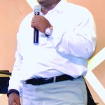 Man speaking on stage with a microphone, wearing a white shirt and glasses.