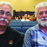 Two men with glasses seated in an auditorium, surrounded by attendees at an event.