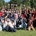 Group of people celebrating outdoors, waving hands, with trees and grass in the background.