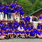 Group photo of young people in matching shirts gathered around a fire truck at a summer camp.
