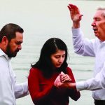 People participating in a baptism ceremony by a lake, two men guiding a woman in red.