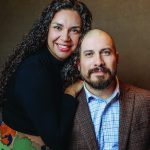 Smiling couple posing together, man in suit with plaid shirt, woman with curly hair in black top.